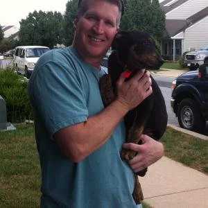 A man holding a dog outside on a sidewalk, with cars and houses in the background.
