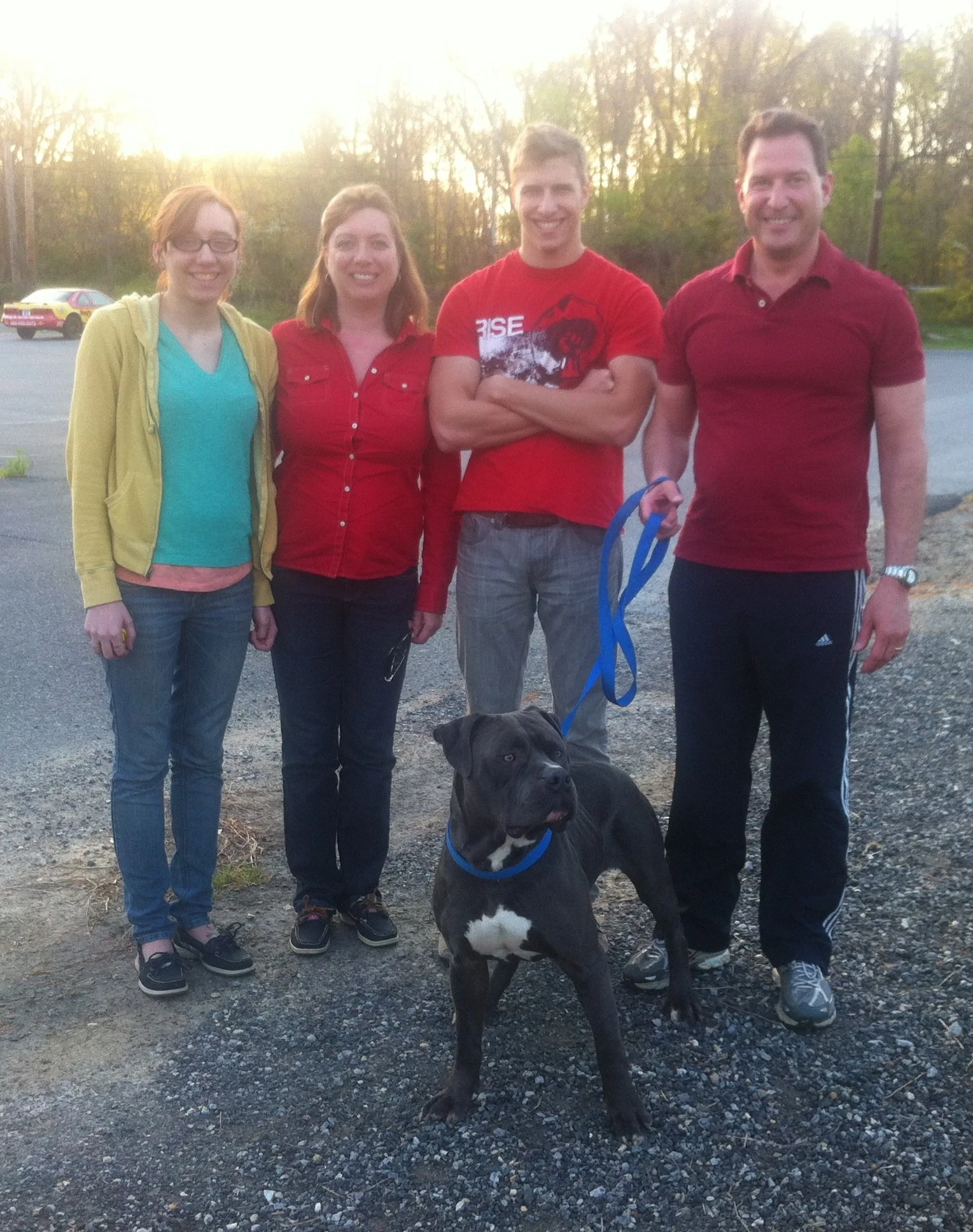 Four people standing outdoors on gravel ground with a large black and white dog, trees, and parked cars in the background. It is sunset or sunrise.