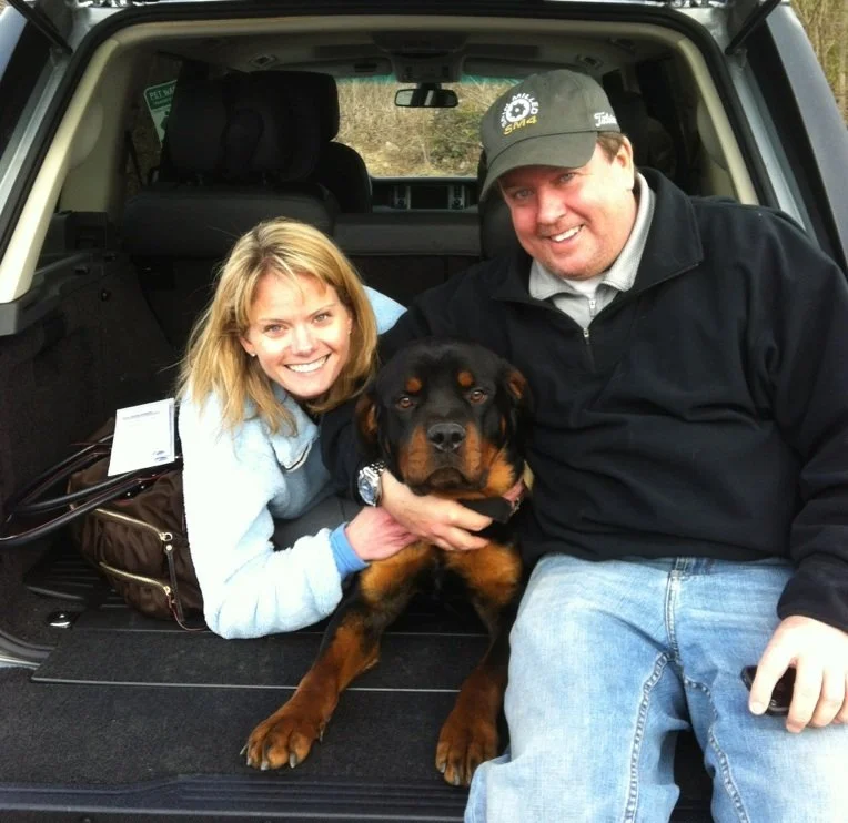 A woman and a man sitting in the back of a vehicle with a Rottweiler puppy between them, all smiling.
