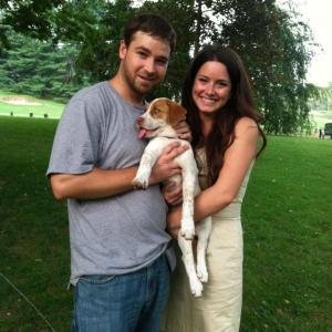 A man and a woman smiling and holding a small dog outdoors in a park.