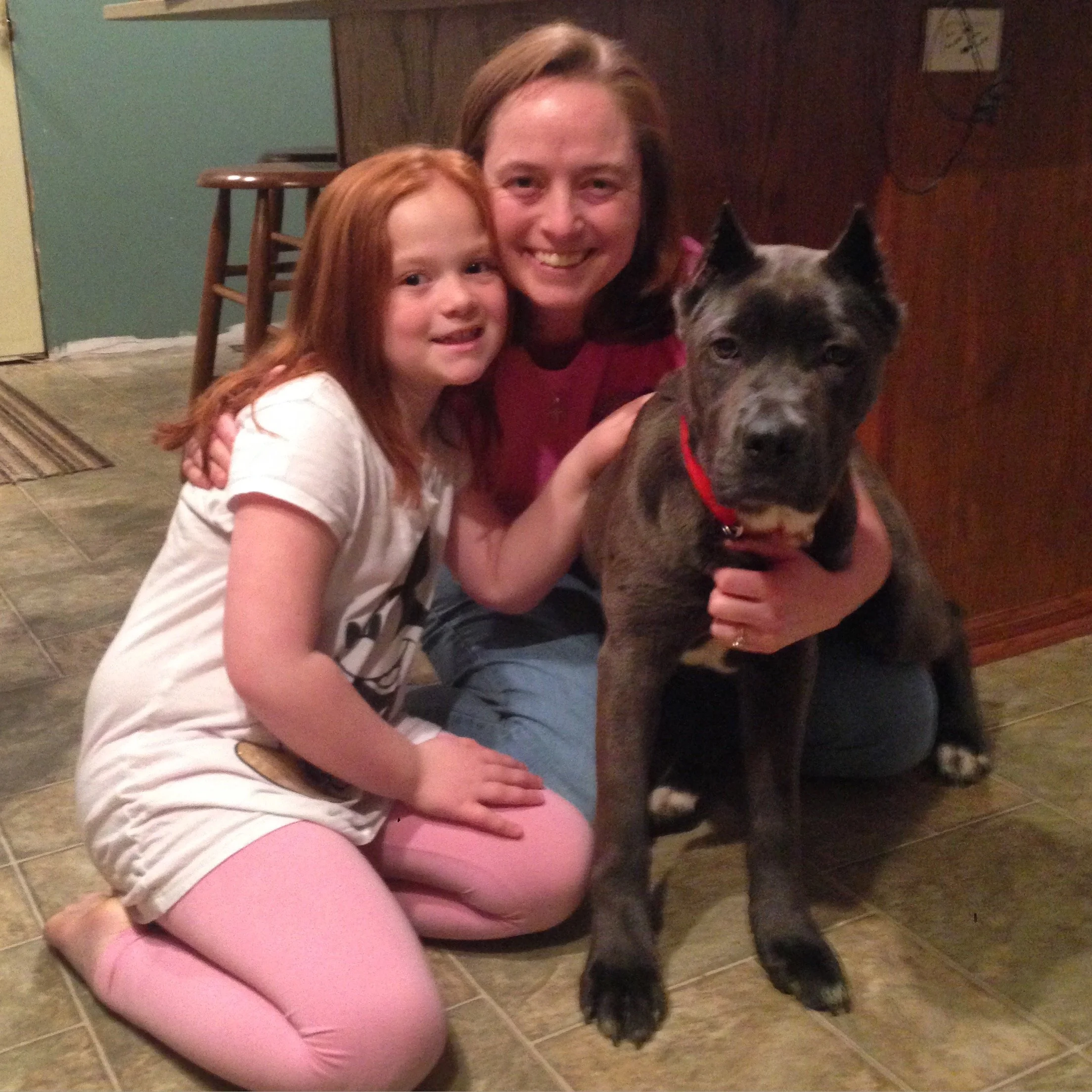 A woman, a young girl, and a dog sitting on a tiled floor inside a house, with the woman and girl smiling and the girl hugging the dog.