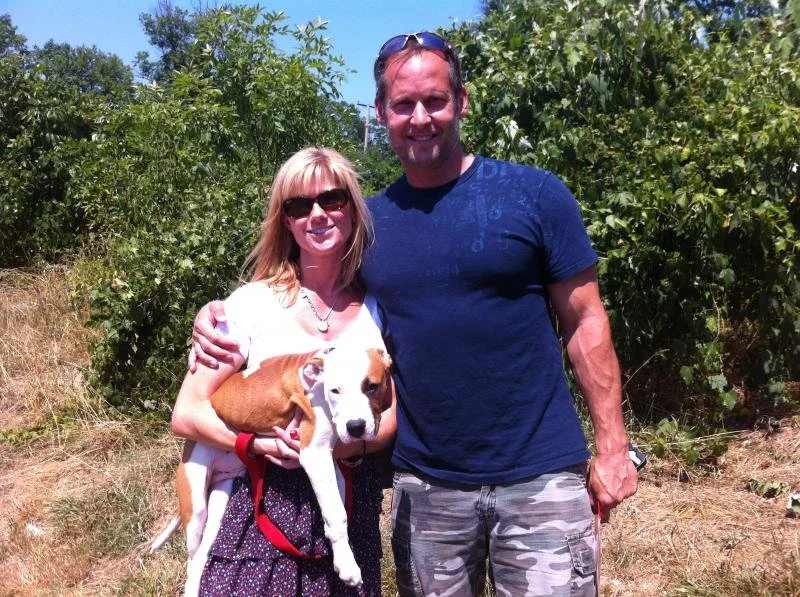 A woman and a man standing outside on a sunny day, smiling. The woman is holding a dog. Green trees are in the background.
