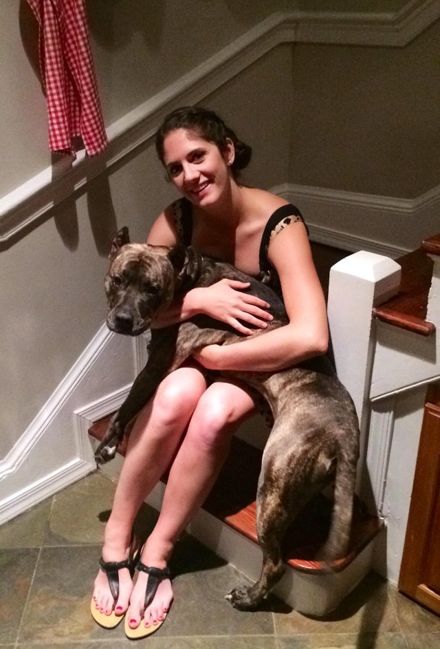 A woman sitting on a staircase holding a large brindle-coated dog in her lap, smiling at the camera.
