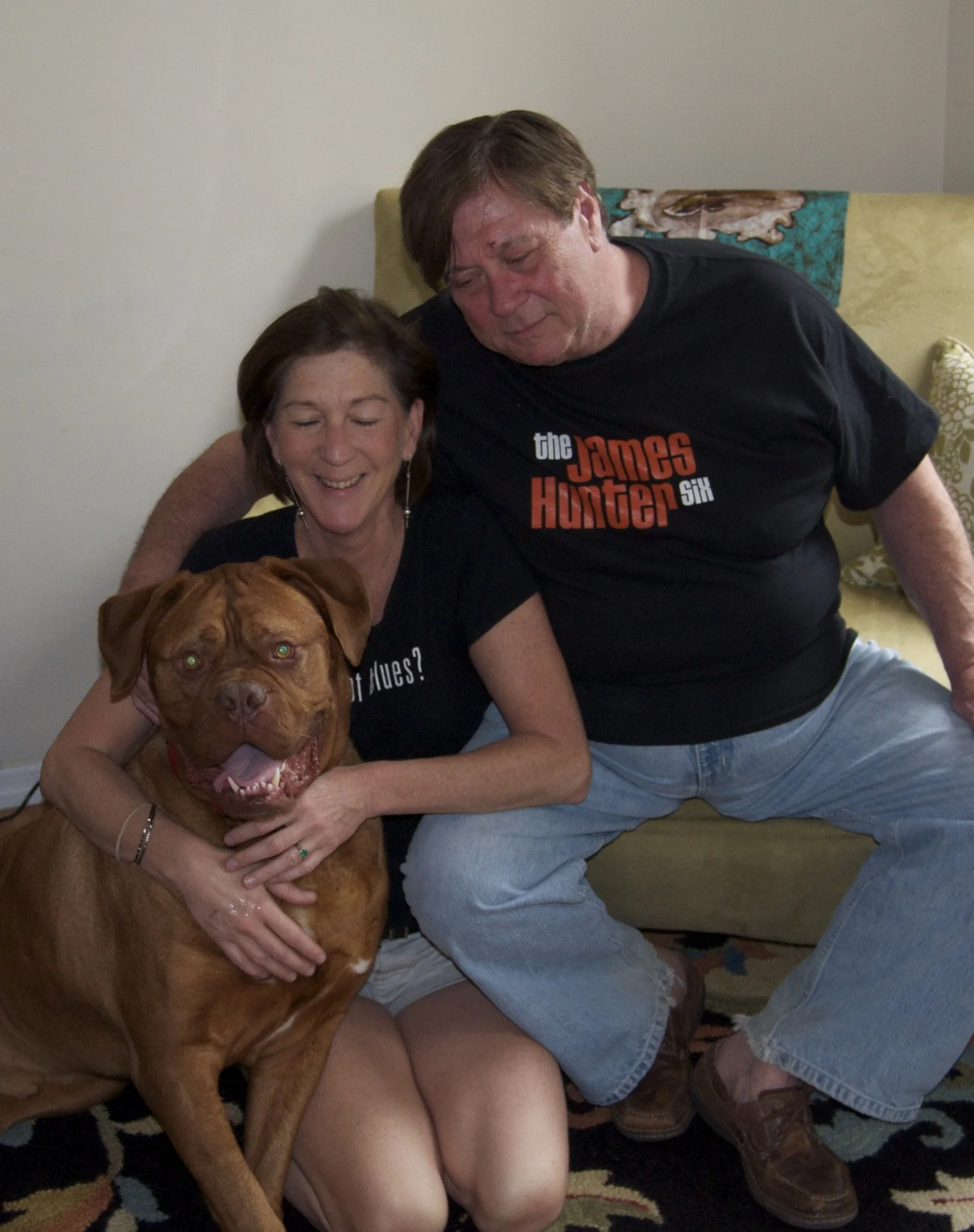 A couple sitting on a couch with a large brown dog. The woman is smiling and hugging the dog, while the man is leaning over her and looking down at the dog. The woman has short brown hair, and the man has short brown hair and is wearing a black T-shi