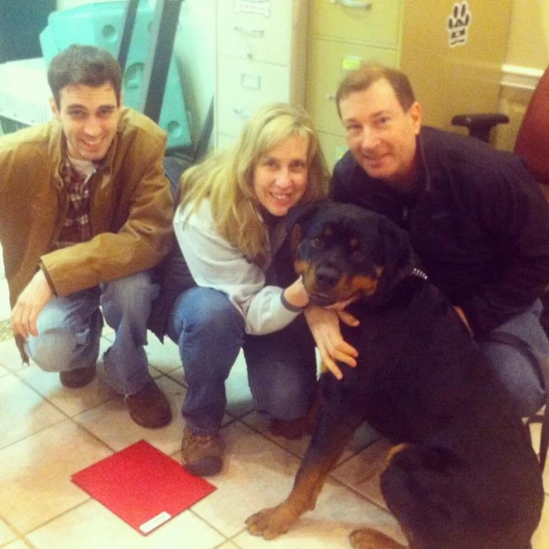 A group of three people and a Rottweiler dog inside a room with lockers and a filing cabinet, posing for a photo. The group appears happy.