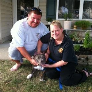 Two people, a man and a woman, crouching on grass outdoors holding a small dark animal, possibly a ferret or similar creature, with a house and potted plants in the background.