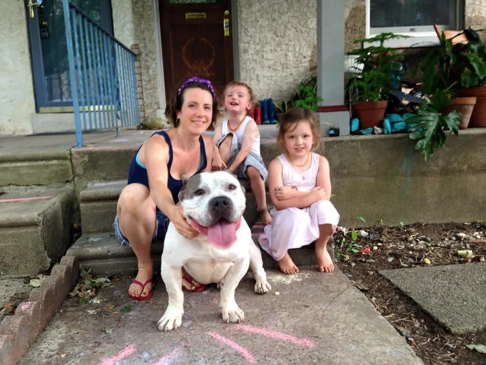 A woman with two young girls and a large white dog outside a house. The woman is kneeling next to the dog, smiling, with her hand on the dog's head. The dog is sitting, with its tongue out. The two girls are sitting on the concrete steps behind them,