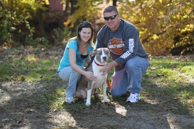 A young woman and a man kneeling next to a large dog outdoors during autumn, with trees in the background showing fall foliage.