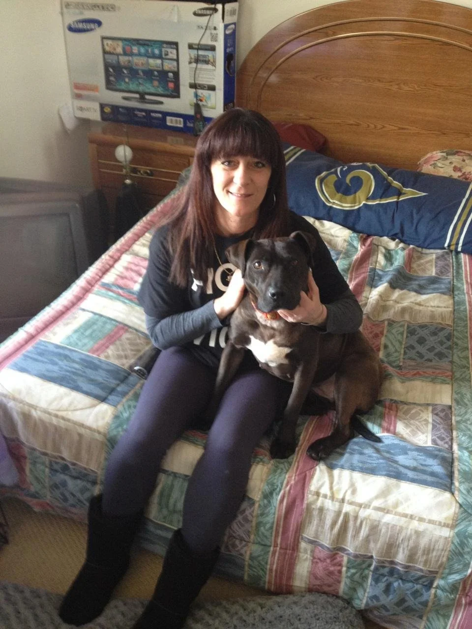 A woman sitting on a bed holding a black dog with a white patch on its chest. There is a large flat-screen TV in a box behind her and a wooden headboard. The bed has a colorful, patchwork quilt.