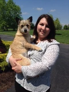 Woman holding a small tan and black dog outdoors in a park with green grass and trees