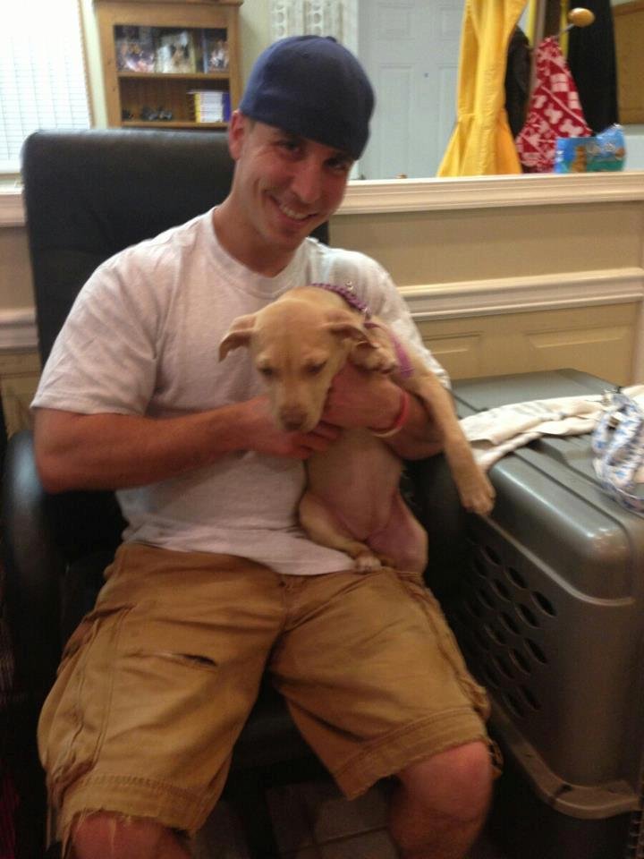 A young man with a blue cap, white t-shirt, and tan shorts sitting in a black chair, holding a small tan puppy, smiling at the camera.