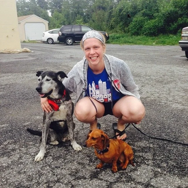 A woman squatting on a parking lot with two dogs, smiling at the camera. The larger dog is a black and gray husky mix with a red bandana, sitting next to her. The smaller dog is a brown dachshund puppy, standing on the ground. There are cars and a ga