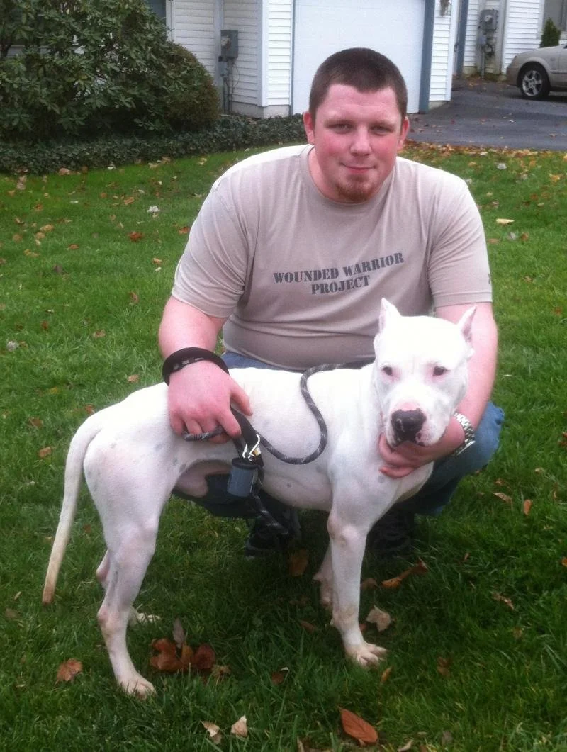 A man kneeling on green grass in front of a house with a grey garage door, holding a white pitbull with a black collar and leash. The man wears a beige T-shirt with the words 'Wounded Warrior Project'.