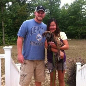 A man and woman standing outside, smiling at the camera, holding a small dog between them, with trees and a white fence in the background.