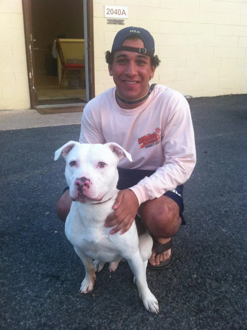 A young man wearing a baseball cap and shorts kneeling next to a white dog with pink spots on its nose, in front of a building with the room number 2040A.