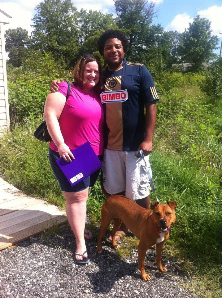 A woman in a pink shirt and a man in a navy and gold shirt stand outdoors on a gravel path, smiling, with their arms around each other. The woman is holding a purple folder, and the man is holding a leash attached to a brown dog. The background featu