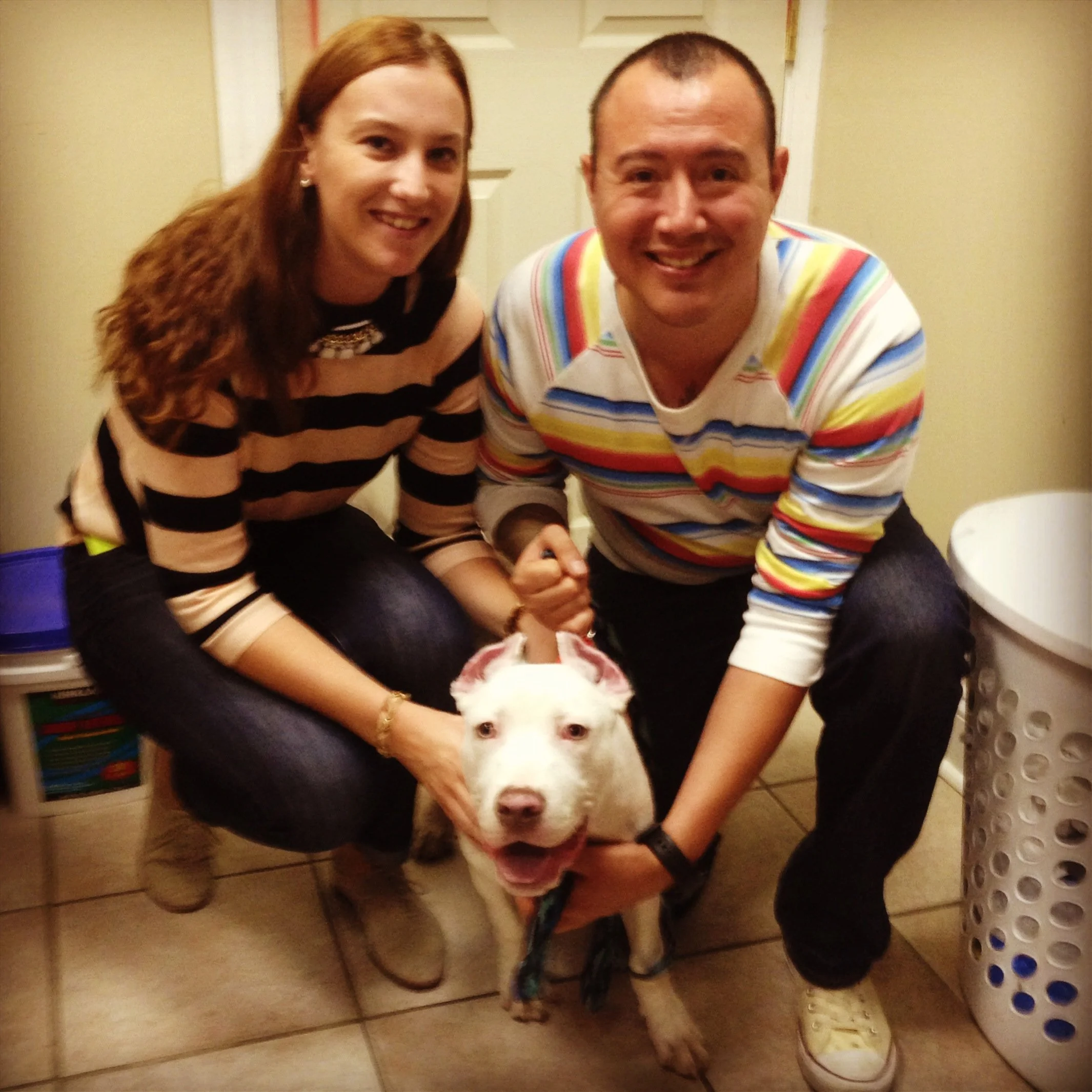 Two people, a woman with red hair and a man with short hair, are posing for a photo. They are indoors with a door and kitchen cabinets in the background, and a white dog with black markings is in the foreground, partially obscured.