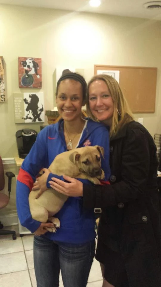 Two women smiling and hugging a light brown dog in an indoor room.