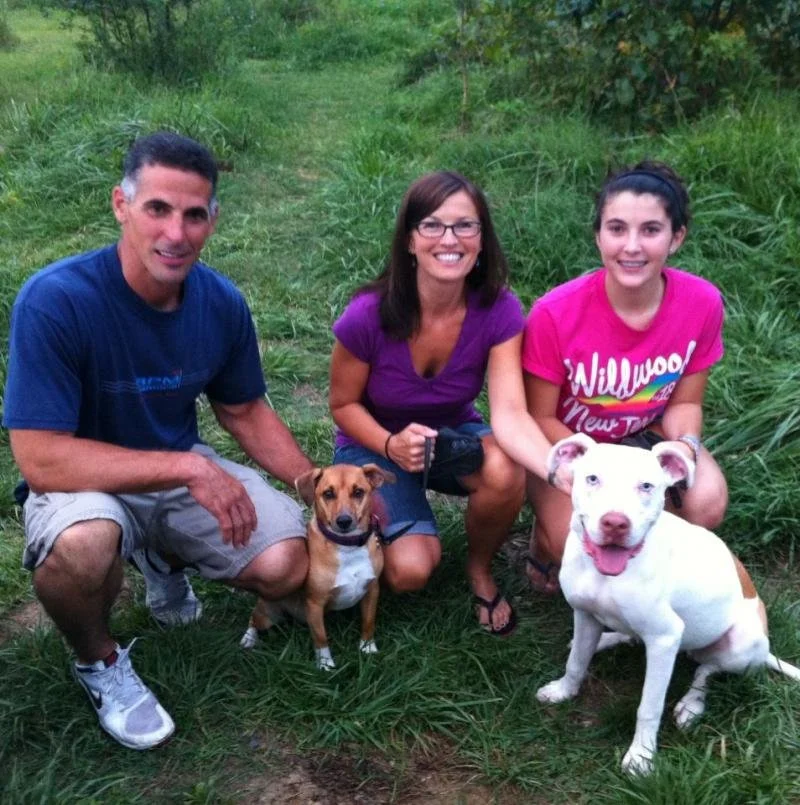 Three people and two dogs outdoors on a grassy path in a natural setting.