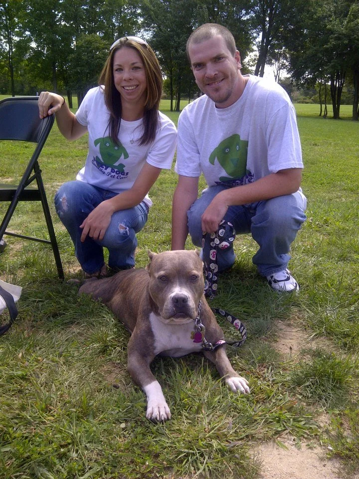 A smiling woman and man crouching on grass outdoors with a dog lying in front of them. The woman has shoulder-length brown hair and is wearing a white T-shirt and ripped jeans, while the man has short hair, a beard, and is wearing a white T-shirt and