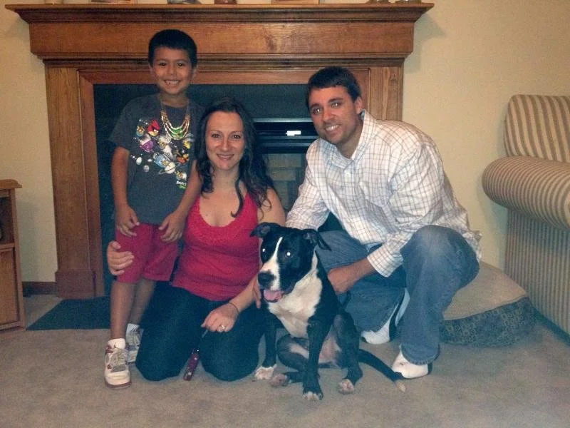 A smiling family of three with their black and white dog sitting on the floor in front of a fireplace in a cozy living room.