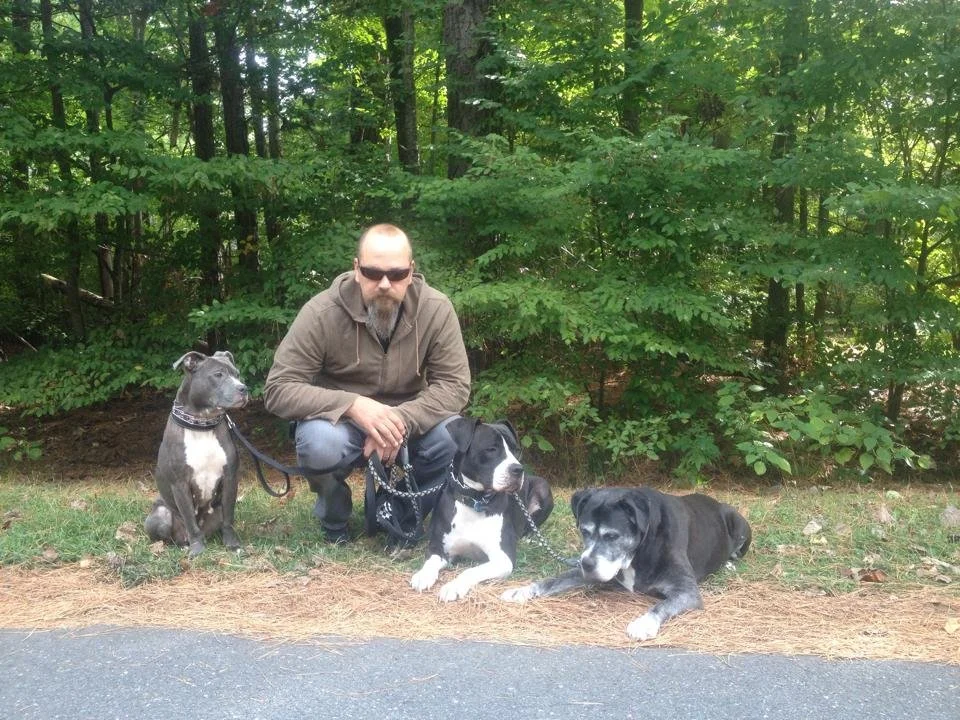 A man wearing sunglasses kneeling outdoors with three dogs near a wooded area.