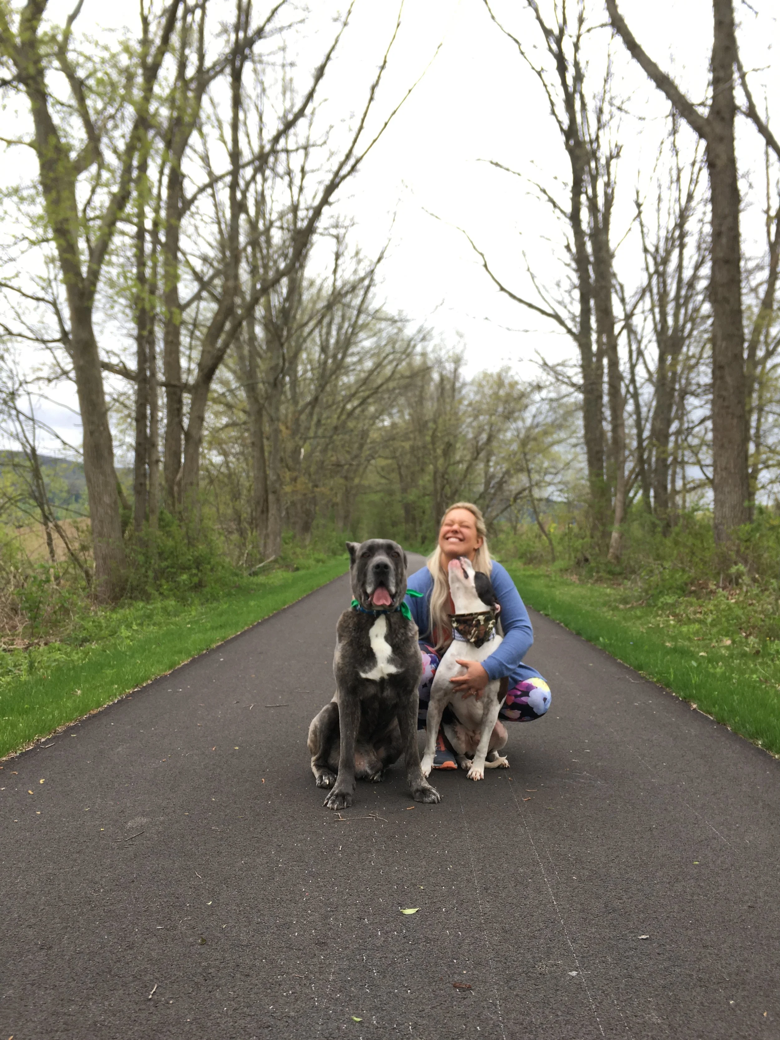 A woman hugging a black and white dog and a woman hugging a gray and white dog on a paved walking trail surrounded by trees.