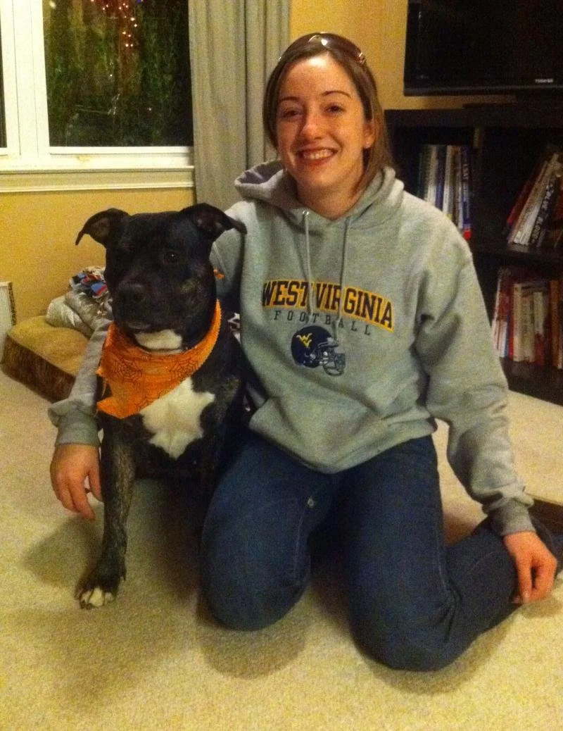A smiling young woman wearing a gray West Virginia football hoodie kneeling on the floor next to a black and white dog with an orange bandana.