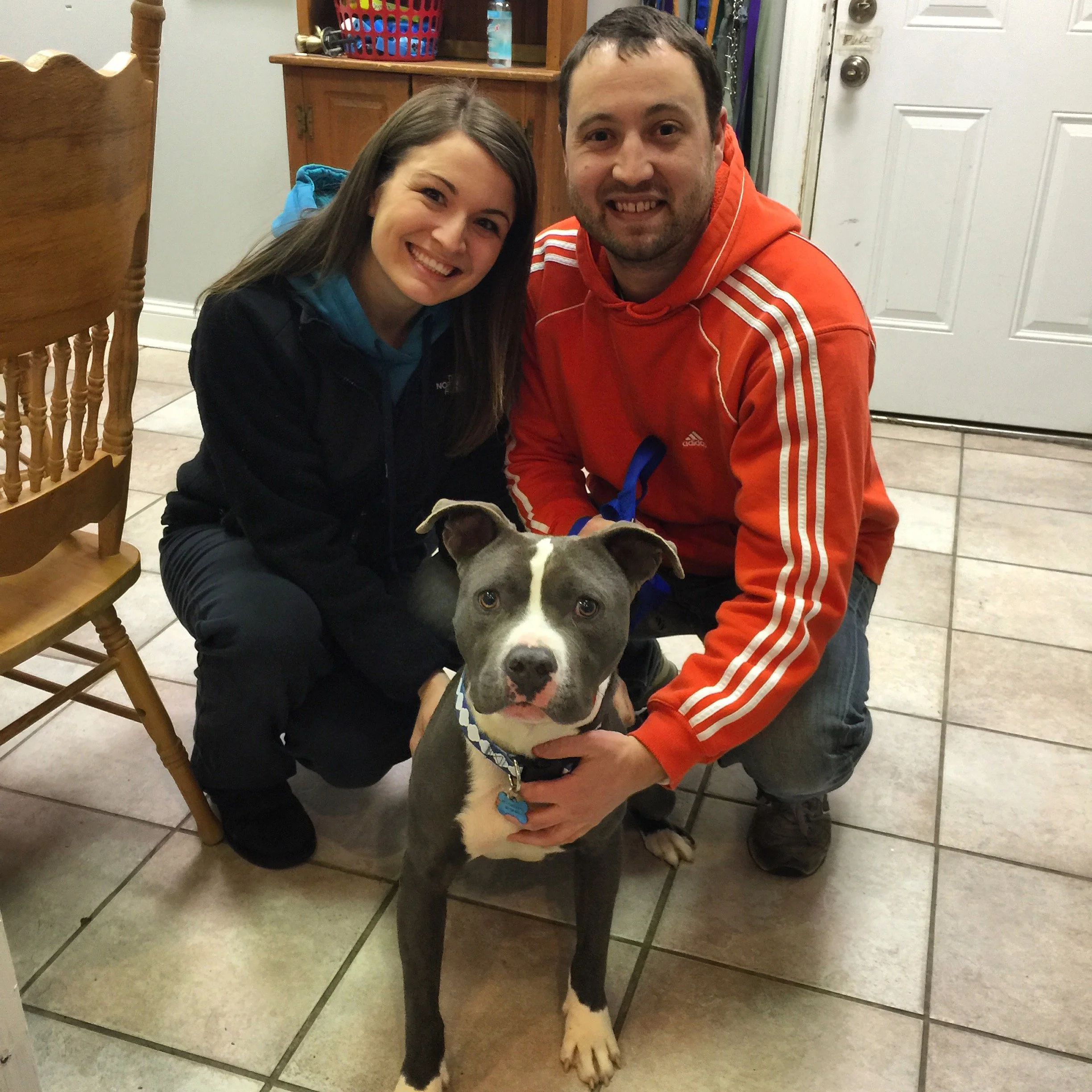 A smiling woman and a man kneeling beside a large dog inside a home with tiled flooring and a white door in the background.