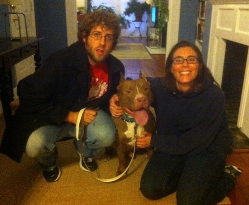 A man and woman with a brown and white dog inside a home, with the dog sitting between them.