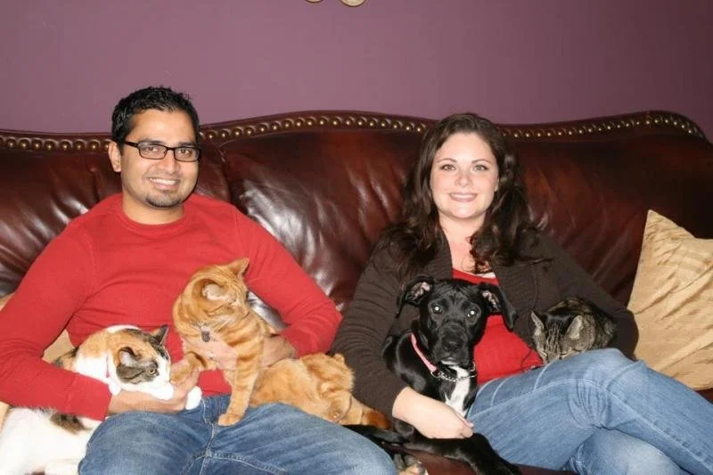 A couple sitting on a leather sofa with their three cats and two dogs, smiling at the camera.