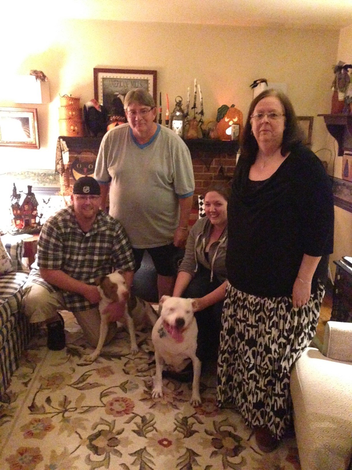 Four people in a living room with two dogs, with various decorations on the wall and mantelpiece.