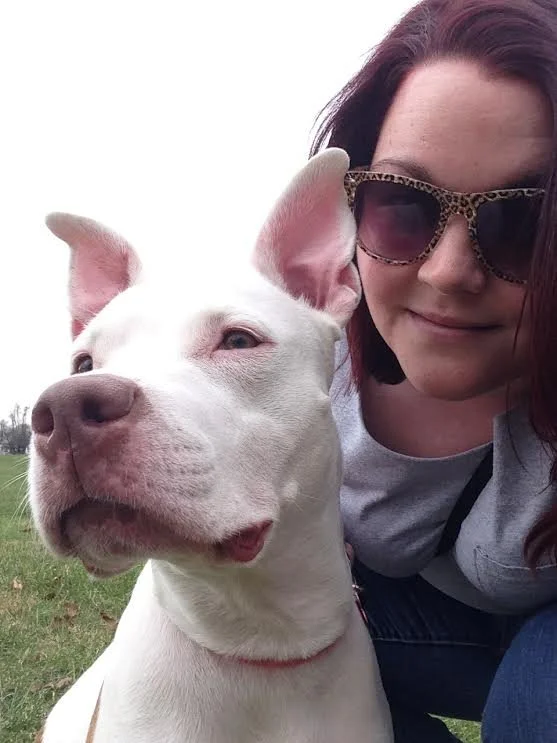 A woman with sunglasses taking a selfie with her white dog outdoors in a park.