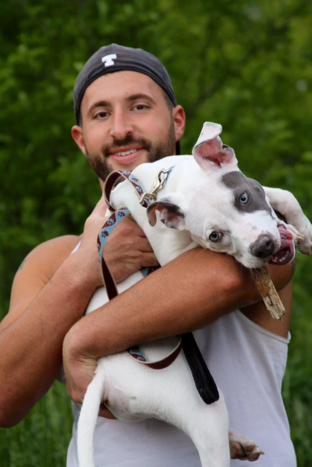 A man with a beard and a black backward cap holding a playful white and gray dog with blue eyes, outdoors with green foliage in the background.