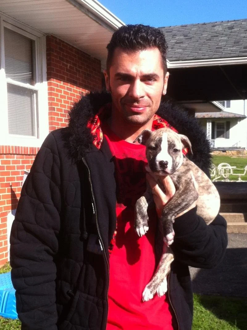 A man holding a gray and white puppy outside a house with a brick wall and a window.