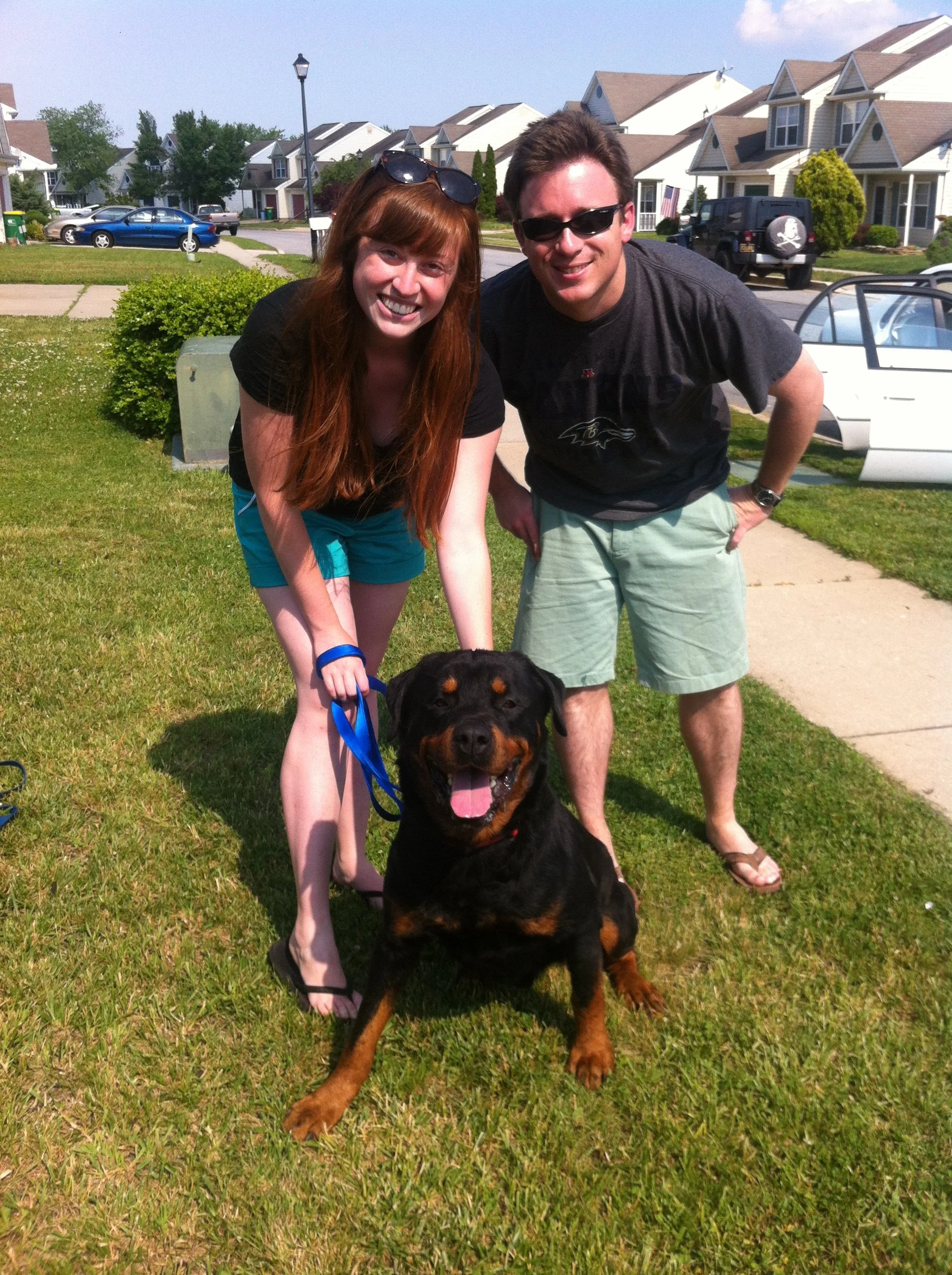 A smiling young woman with long red hair and a young man with short brown hair, both wearing sunglasses, posing outdoors on a suburban street with a large Rottweiler dog sitting in front of them.