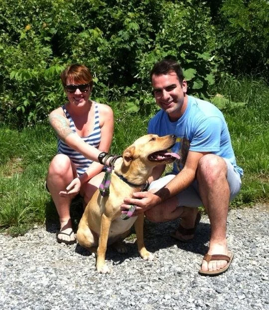 A woman and a man kneeling on a gravel path outdoors with a tan-colored dog between them. The woman has short hair, sunglasses, a striped tank top, and tattoos on her arm. The man has dark hair, is wearing a blue t-shirt, and sandals. They are smilin