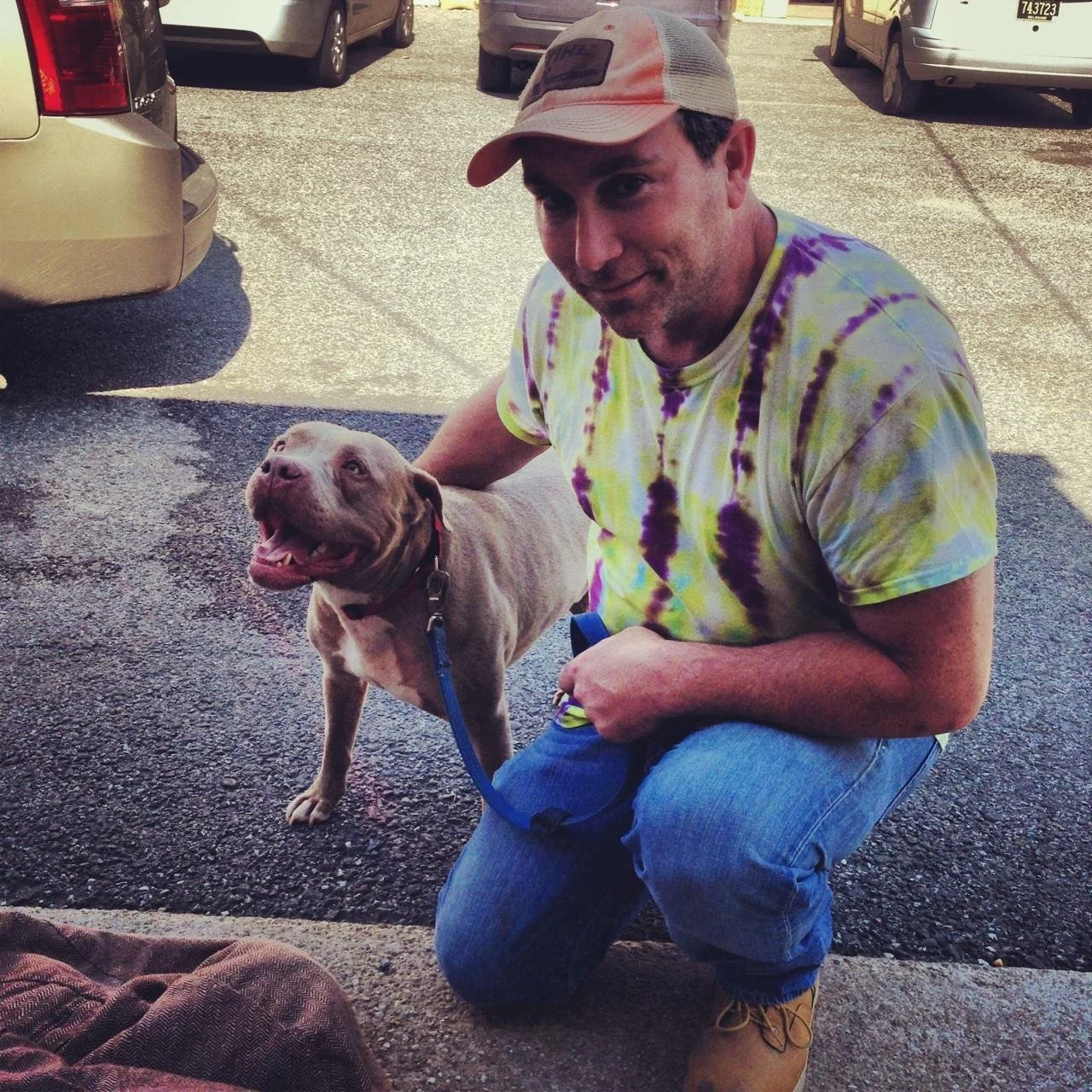 A man wearing a colorful tie-dye T-shirt, blue jeans, and a trucker hat kneels on one knee beside a smiling, brown pitbull dog on a leash in a parking lot.