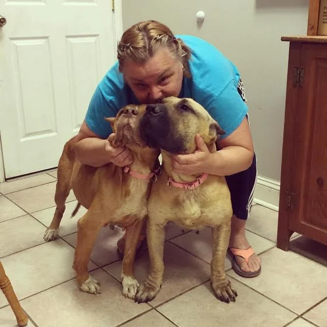Woman hugging two large dogs in a room with tiled floor and wooden furniture.