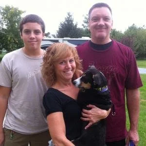 A woman with curly red hair holding a black dog, and two young men standing beside her outdoors with trees and grass in the background.