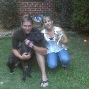 Two people kneeling on grass outdoors, each holding a small dog, with a brick wall and bushes in the background.