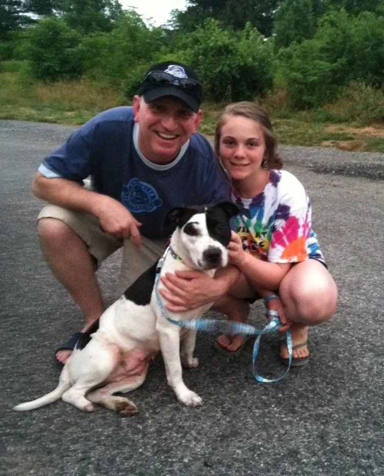 A man and a girl squatting on a gravel road, smiling, while holding a black and white dog. They are outdoors with green trees in the background.