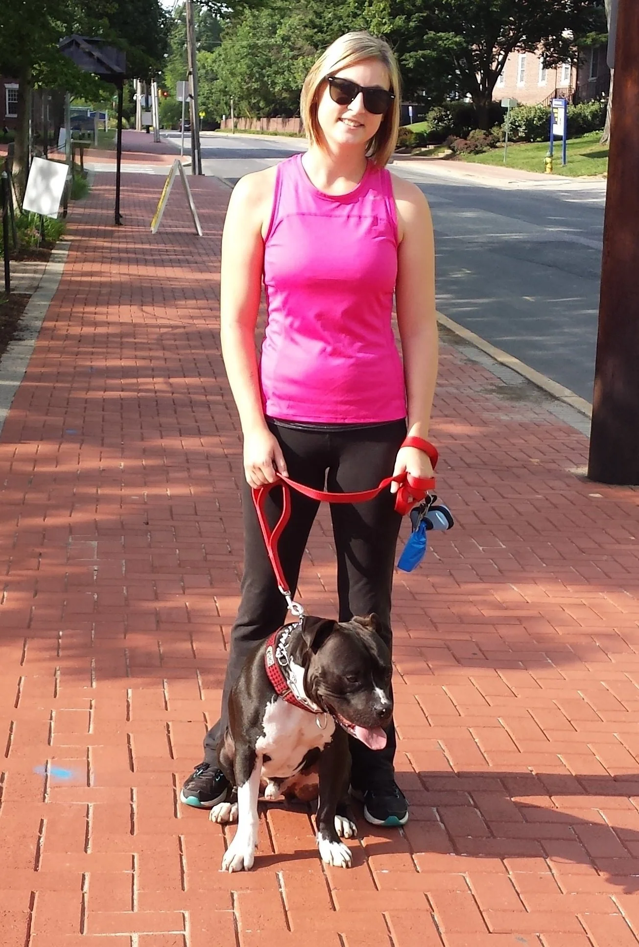 A woman in a pink athletic top and black pants holding a red leash with a dog walking on a brick sidewalk in a small-town street scene.