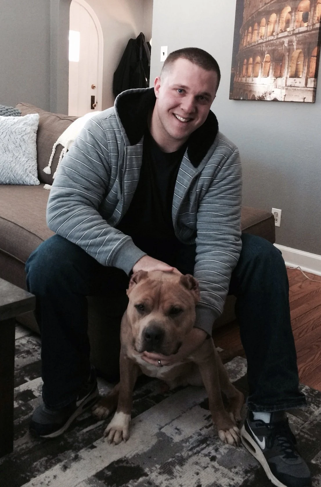 A young man with short hair, wearing a gray hoodie with black lining, sitting on a couch and smiling at the camera. He has a brown dog with white markings sitting in front of him on a patterned rug in a living room.