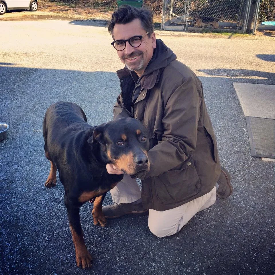 A man kneeling on the ground next to a large black and tan dog outdoors, with a fenced area in the background.