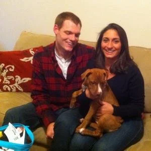 A young couple sitting on a couch holding a brown puppy, smiling at the camera.