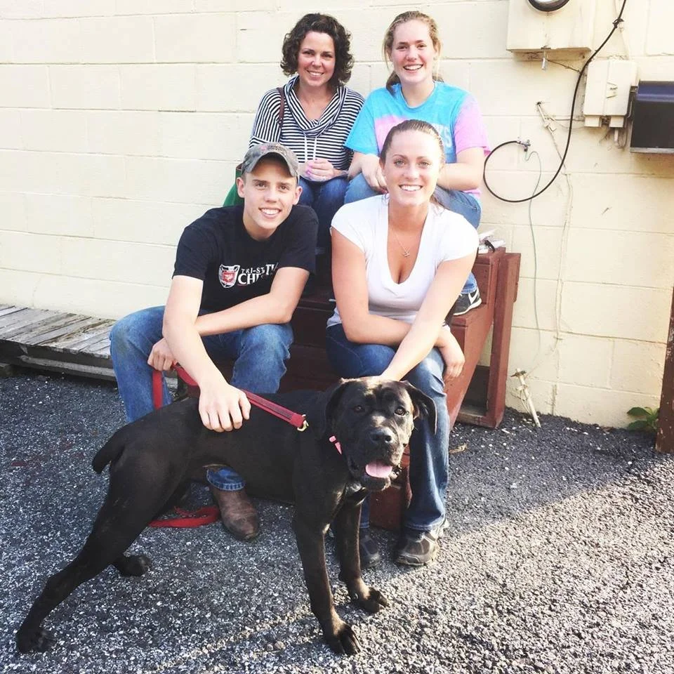 Four people and a dog posing outdoors in front of a beige wall. The group includes two women and two teenagers, all smiling. The woman at the front right is crouching, and a large black dog with a red leash is standing beside her, looking at the came