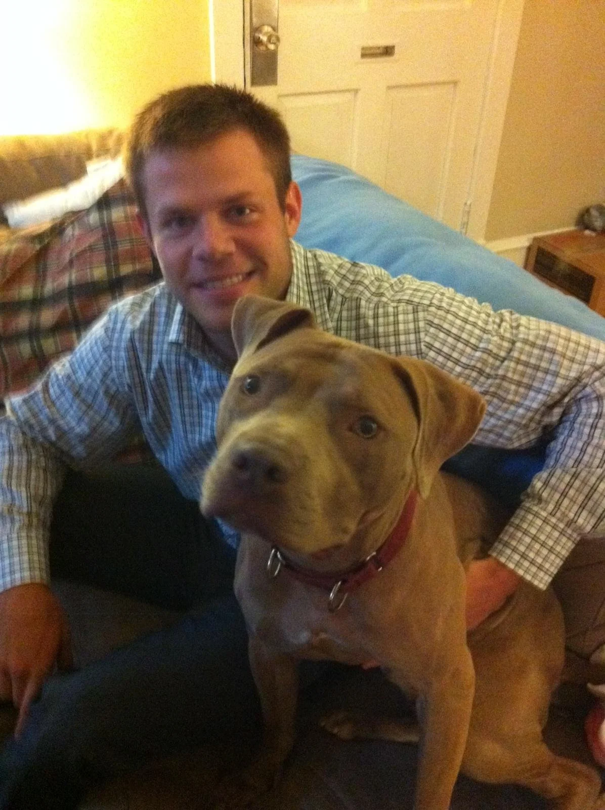 A man smiling and sitting on a couch with a large, tan pitbull dog in front of him. The man is wearing a striped shirt and the dog is wearing a red collar.