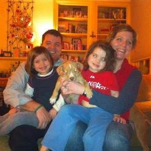 A family of four with two young girls and an adult man and woman sitting together in a warmly lit living room, holding a small dog.
