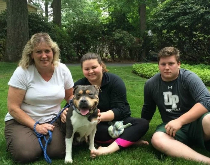 Four people and a dog sitting on grass in a backyard with green trees and bushes in the background.